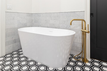 A freestanding tub with a gold faucet, black and white pattern tile flooring, marble subway tile...
