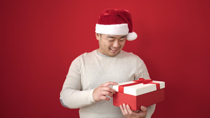 Young chinese man wearing christmas hat holding gift over isolated red background