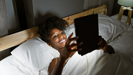 African american woman using touchpad and earphones lying on bed smiling at bedroom - obrazy, fototapety, plakaty