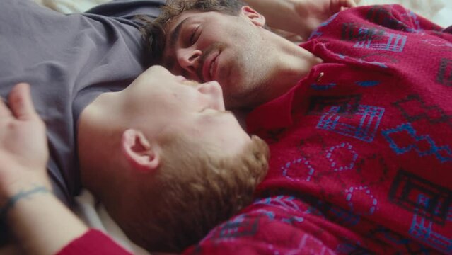 Close-up view of handsome gay couple resting on bed at home, talking to each other, kissing and smiling