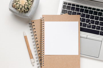 Paper blank mockup card on notepad. Workspace with paper card, laptop, coffee, office supplies and cactus plant on white wooden table. Home office, aesthetic style, top view.