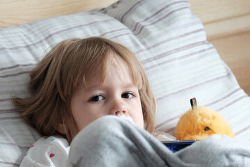 A small fair-haired, European-looking girl of two years old lies in bed with a pacifier, looks,...