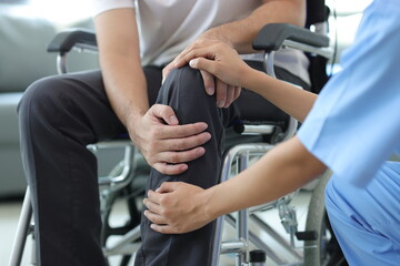 Female doctor diagnosing knee pain of male patient in wheelchair in hospital examination room. - obrazy, fototapety, plakaty