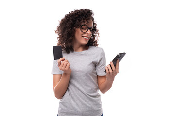 Naklejka premium cute slim curly woman with black hair dressed in casual gray t-shirt holding smartphone and plastic credit card