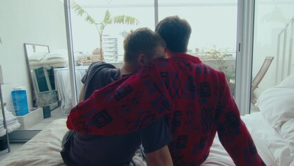 Romantic gay couple sitting on bed at home, looking at cityscape through the window, embracing and kissing each other with love. View from behind, zoom shot