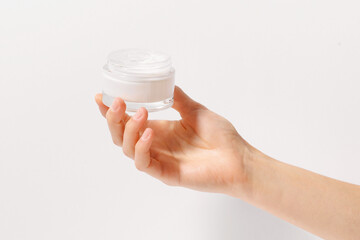 Close-up of a female hand holding a transparent glass jar of white face cream on a white isolated background. Concept of beauty products, daily hygiene and skin hydration