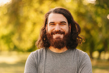 Close up portrait of bearded hipster man with long hair looking at smiling at the camera whie standing outdoor in park.