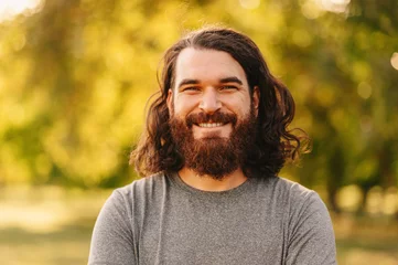 Fotobehang Close up portrait of bearded hipster man with long hair looking at smiling at the camera whie standing outdoor in park. © Vulp