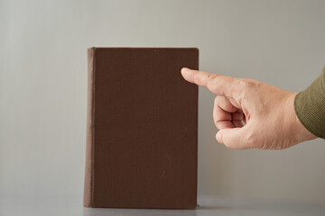 A hand with a pointing finger pointing at an old book with no title on the cover. Law of morality, old testament, old laws of morality, divine law - obrazy, fototapety, plakaty