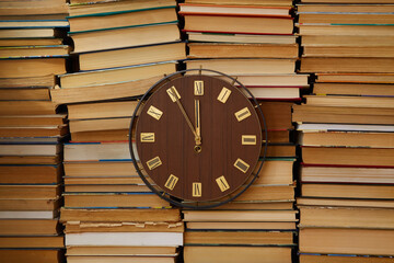 Large clock with Roman numerals on the clock face against a backdrop of stacked books. Old-school,...