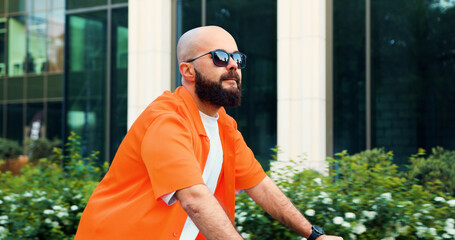 Happy bearded man in casual clothes and white casual headphone riding bicycle at urban street. Professional young male going to work on bicycle in city street.