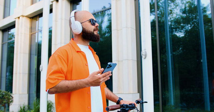 Happy bearded caucasian bearded man in headphone and sunglasses using smartphone while walking with bike at urban area. Commute work sustainable lifestyle in the city. - Powered by Adobe