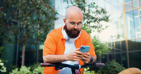 Positive man with bicycle wearing orange shirt texting on phone and looking around near building...