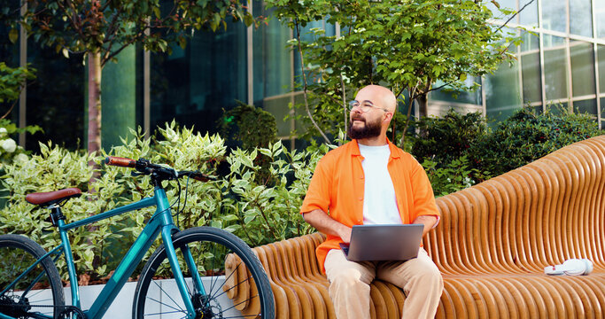 Cheerful smiling bearded man in glasses typing on keyboard sitting on bench with his bicycle beside him outdoor. Handsome guy working on laptop computer near modern glass office.