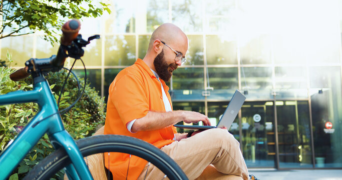 Stylish hipster man in glasses working in laptop while sitting in city square on bench with bicycle at sunset. Freelancing and outdoor education concept.