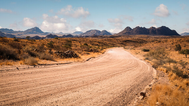 View of the Namib Desert in northern Erongo, Namibia, with mountains in the background and a car driving on a  gravel road  kicking up dust behind it.