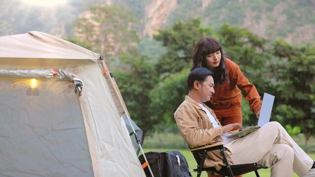 Couple relaxing at camping tent in nature, planning route for leisure travel, searching information on laptop. Mountain valley during sunset. Natural summer landscape on background.