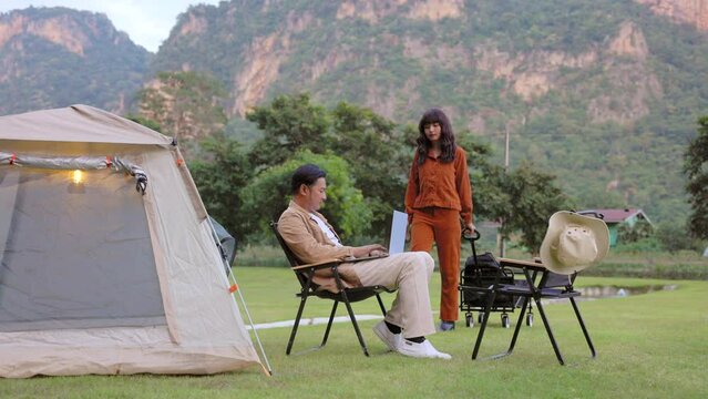 Young beautiful couple relaxing at camping area in evening. Asian woman dragging camping cart and looking at laptop together, shop on internet, choose product to order online, watch streaming service.