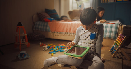 Portrait of Little Korean Boy in his Room Playing Video Games on a Tablet Computer While his Sister is Reading a Book in the Background. Happy Children in Pyjamas Spending Their Weekend at Home