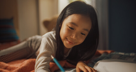 Portrait of a Little South Korean Girl Drawing with Colored Pencils in a Notebook While Lying in Bed in Children's Bedroom at Home. Adorable Asian Kid Playing in Her Cozy Room with Toys