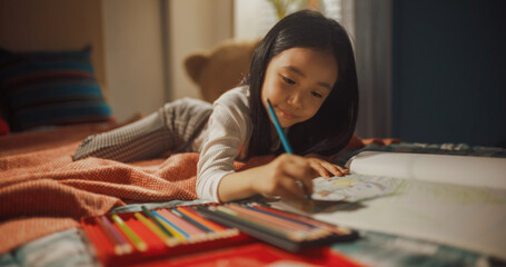 Portrait of a Little South Korean Girl Drawing with Colored Pencils in a Notebook While Lying in...