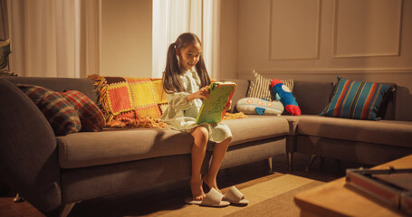 Wide Shot Portrait of a Little Korean Girl Sitting on a Couch in the Living Room and Using Tablet Computer to Play Video Games. Active Female Child Using Interactive Media for Learning
