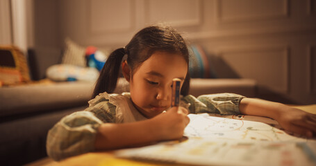 Close Up Portrait of a Little Cute Korean Girl Drawing in her Coloring Book in a Living Room at Home. Smart and Creative Female Child Spending her Evening Learning, Studying, Doing Homework