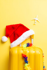 Island-bound for New Year’s! Top-view vertical photo featuring suitcase, Santa hat, floral garland, and a miniature plane on a vibrant yellow backdrop. Get ready for a tropical celebration