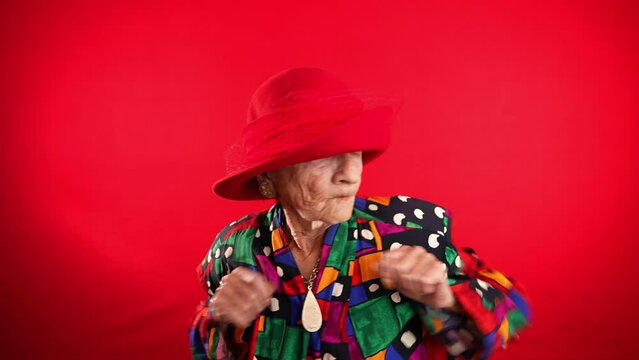Funny fisheye portrait of elderly woman doing Dab dance wearing red hat on red background.