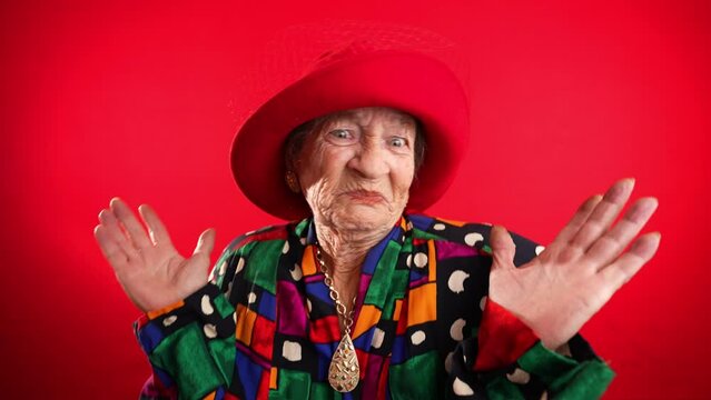 Portrait of concerned, unhappy elderly senior old funny crazy woman with wrinkled skin red hat and no teeth put hands out ask WHY ME, isolated on red background studio.