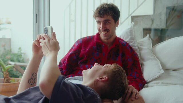 Young gay man lying on bed and discussing social media post on smartphone with boyfriend while bonding together at home