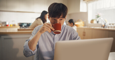 Portrait of Korean Family at Home: Young Father Using Laptop Computer for Working Remotely, With his Wife and Kids Preparing Lunch in the Background. Supportive Family of a Businessman