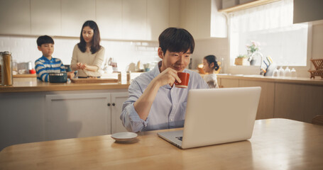 Korean Family at Home: Young Father Using Laptop Computer for Working Remotely While Waiting for Food, With his Wife and Kids Preparing Lunch in the Background. Supportive Family of a Businessman - obrazy, fototapety, plakaty