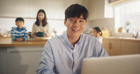 Portrait of Young Korean Man Working From Home on Laptop Computer, Looking at the Camera and Smiling with his Loving Family in the Background. Successful Businessman Having the Support of Loved Ones