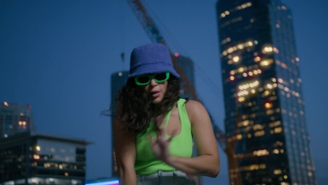 Cool girl dancer dancing in night city. Black young girl with long curly hair moves rhythmically. Sexy fashionable dancer in trendy bucket cup and bright green glasses moves sensually urban downtown