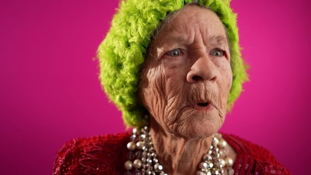 Unhappy displeased fisheye view caricature of funny elderly woman saying NO with green wig or hat isolated on pink background in slow motion.
