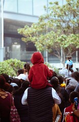 Vertical back view of people gathered, a kid with a red jacket sitting on the shoulders of a male