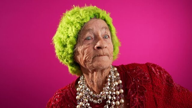 Funny fisheye portrait of elderly woman doing Dab dance wearing green wig or hat isolated on pink background in slow motion
