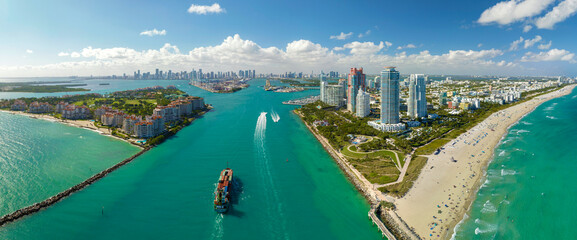 Commercial container ship entering Miami port harbor through main channel near South Beach. Luxurious hotels and residential buildings on waterfront and high skyscraper towers of downtown in distance - obrazy, fototapety, plakaty