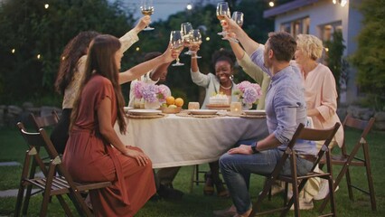 Group of diverse people celebrating, chilling out at home party outdoors at garden at home. Big family and neighbor gathering enjoy dinner and clicking glasses of alcohol together at yard front home.