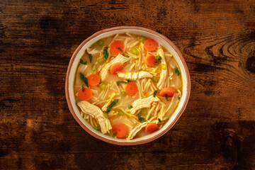 Chicken noodle soup with vegetables, a bowl of healthy broth, overhead flat lay shot on a rustic wooden table, winter comfort food