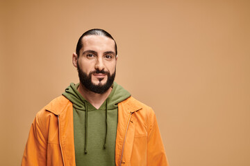 portrait of handsome and bearded arabic man in casual attire looking at camera on beige backdrop
