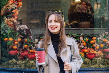 Portrait of young smiling woman in coat enjoying drink in reusable coffee cup walking on street...