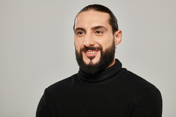 portrait of cheerful and handsome arabic man with beard posing in black turtleneck on grey backdrop