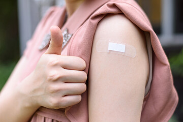 Vaccinated Woman Gesturing Thumb Up and Showing Arm With Plaster Bandage After Coronavirus Vaccine Injection. Covid-19 Vaccination concept. - obrazy, fototapety, plakaty