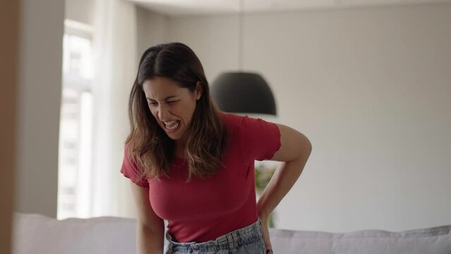 Caucasian woman standing up from a couch at home experiences sudden back pain. Young adult female suffers from pain in her back raising from sofa. Girl rubbing back suffering from cramps in lumbar
