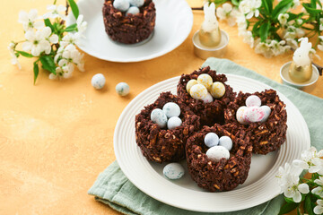 Easter chocolate nest cake with mini chocolate candy eggs with blossoming cherry or apple flowers on yellow background table. Creative recipe for Easter table with holiday decorations. Top view.