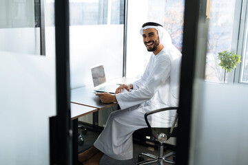 Handsome man with dish dasha working in his business office of Dubai. Portraits of a successful businessman in traditional emirates white dress. Concept about middle eastern cultures.
