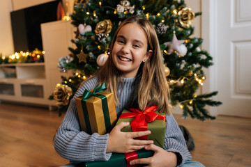 Portrait of cute little girl in sweater holding with many Christmas gifts boxes sitting on background of xmas tree, smiling looking at camera with happy expression. Concept of home festive atmosphere.