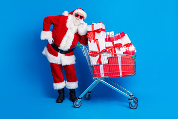 Full body photo of aged senior man lean on shopping trolley wear trendy santa claus costume coat isolated on blue color background